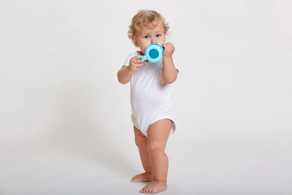 ブロンドの巻き毛の赤ちゃんはボトルから水を飲み カメラを直接見て ボディスーツを着て 子供の完全な長さの写真は白い背景に対してポーズをとって 赤ちゃんカップを保持しています — ストック写真