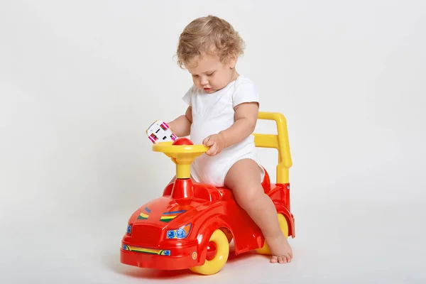 小さな魅力的な赤ちゃんの男の子が赤と黄色のトロカーに座って 手の中におもちゃの車を保持し 白いボディスーツを着て 金髪の巻き毛 かわいい赤ちゃんが屋内で遊んでいる — ストック写真