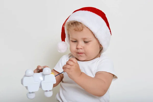 彼の新しいおもちゃ クリスマスプレゼント 手の中に保持しているプラスチック製の犬のおもちゃ それを注意深く見て 白いTシャツとサンタの帽子を身に着けている — ストック写真