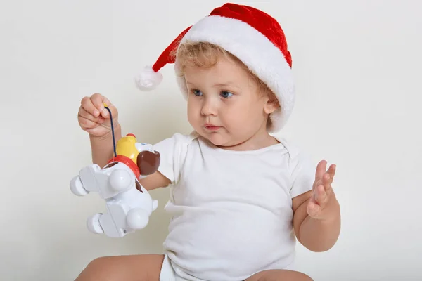 かわいい赤ちゃん身に着けているクリスマスの帽子とボディスーツで遊ぶプラスチック製の犬 魅力的な幼児は彼のおもちゃを保持し 子供は離れて見て 赤ちゃんは離れて見て 新しい興味深いおもちゃを脇に見る — ストック写真