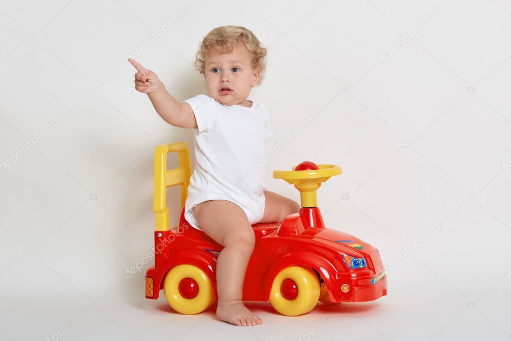 Toddler posing while sitting on toy race car, looking away and indicating with index finger, dresses white bodysuit, baby boy playing indoor, sees something aside, wants to get new toy.