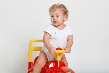 Heyecanlı erkek bebek, kırmızı ve sarı renkli bir arabada oturuyor, ilgiyle başka tarafa bakıyor, beyaz tulum giyiyor, yeni oyuncağıyla oynuyor, arka planda izole bir şekilde..