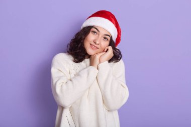 Noel Baba şapkası ve beyaz kazak giyen, doğrudan kameraya bakan, avuçlarını yanaklarına yakın tutan, eflatun arkaplan üzerinde izole edilmiş, sakin ve şirin bir yüz ifadesi olan hoş bir kadın portresi..