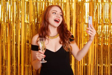 Sarı arka planda yalnız poz veren güzel bir kadın. Süslerle süslenmiş. Bir bardak şarapla gülümseyen genç bir kadın akıllı telefondan selfie çekiyor. Siyah elbiseli ve kolyeli, heyecanlı kızıl saçlı bir kız..
