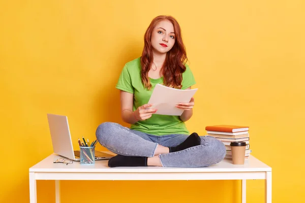 图片中可爱的年轻女性在工作或学习时坐在笔记本电脑和书本旁边 坐在桌子上 双腿交叉 严肃地看着镜头 靠着黄色的墙壁摆姿势 — 图库照片
