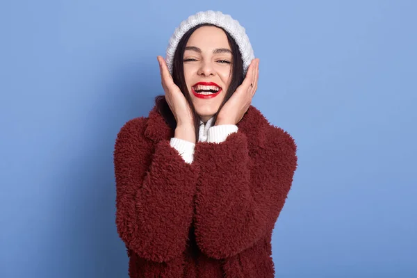 赤い唇を持つ積極的な美しい女性は 頬に両手を保ち 白い冬のセーターと暖かいコートを着て 魅力的な笑顔でカメラを見て 青い壁の上に隔離され 女性は喜んで笑っている — ストック写真