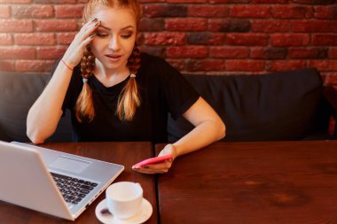 Diz üstü bilgisayar ve akıllı telefon kullanan kızıl saçlı güzel bir kız koltukta şaşkın bir yüz ifadesiyle oturuyor, cep telefonuyla şok edici haberler okuyor, duvara karşı poz veriyor..