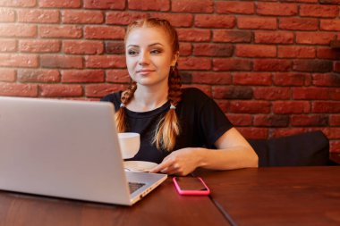 Kafede dizüstü bilgisayar kullanan kızıl saçlı, seramik bardaktan kahve içen, defter ekranına bakan, iki örgülü saçlı çekici kız, duvarın üzerinde izole bir şekilde poz veriyor..