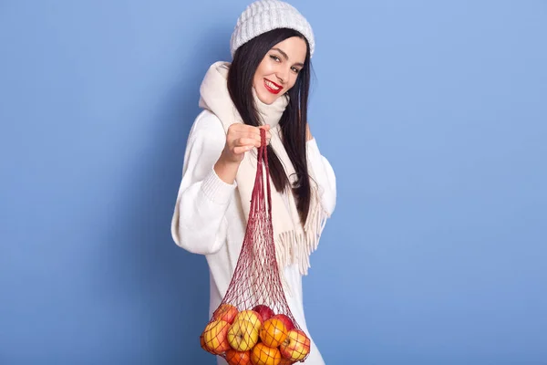 迷人的年轻女子拿着苹果和橘子在网袋里 女性真诚地笑着 满脸满意地看着相机 穿着暖和的毛衣 围巾和帽子的女士 — 图库照片