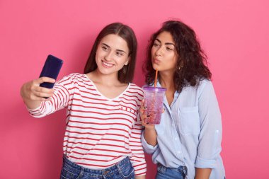 İki mutlu ve çekici kız arkadaş esmer kadın. Biri kokteyl içip telefonda selfie çekiyor. Pembe arka planda soyutlanmış poz veriyor..