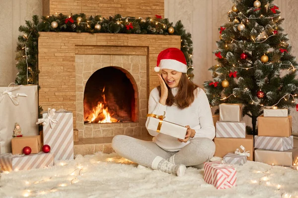 暗い髪を持つ美しい若い女性 暖炉やクリスマスツリーの近くに座っている間 手でお祝いの贈り物を保持 幸せな女の子ドレスでサンタの帽子 暖かいセーターと靴下 — ストック写真