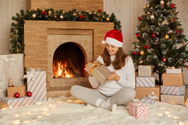 素敵な女性はクリスマスに贈り物を開き 暖炉や木の近くの床に座って サンタの帽子 白いセーター 暖かい靴下を身に着けている女性を笑顔 交差するラグと現在のボックスを保持して座っています — ストック写真