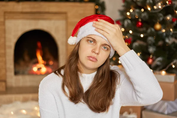 Stressed christmas woman having headache, keeping palm on her forehead, looks sick and unhappy, looking directly at camera, suffering from cephalagia, fireplace and x-mas tee on background.