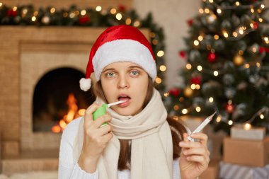 Genç bir kız boğazına ağrıdan fışkıran spreyle ağzına su sıçrıyor. Soğuk algınlığı tedavisi, grip, termometreyi elinde tutmak, kameraya bakmak, Noel tatilinde hasta olmak..