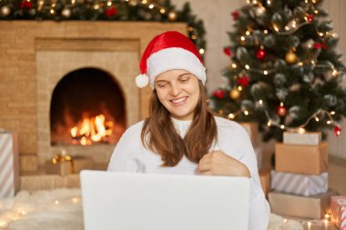 Genç Avrupalı bir kadın Corona virüsü salgını nedeniyle Noel 'i evde kutluyor, yakın bir insanla görüntülü görüşme yapıyor, bilgisayar ekranına gülümsüyor, beyaz kazak giyiyor ve Noel Baba şapkası takıyor..