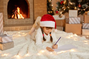 Küçük tatlı bayan çocuk halıya uzanıyor ve Noel Baba 'ya mektup yazıyor, boş kağıda bakıyor ve düşünüyor, şimdiki kutular, Noel ağacı ve yangın yerlerine poz veriyor..