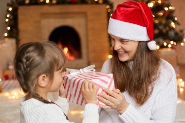 Küçük kızına Noel 'i yaşatan sevimli bir kadın, Noel Baba şapkası takmış, çocuğuna gülümseyen bir kadın, Noel Baba' nın yeni yılını kutlamak için oturma odasında, mutlu bir aile kış tatilini kutluyor.