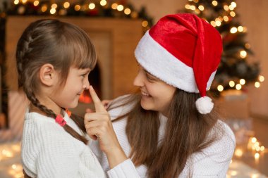 Genç, gülümseyen anne ve küçük kız Noel 'de oynaşıyor, birbirine sevgiyle bakıyor, anne kadın çocuk burnuna dokunuyor, at kuyruklu çocuk utangaç görünüyor, dekore edilmiş oturma odasında poz veriyor..