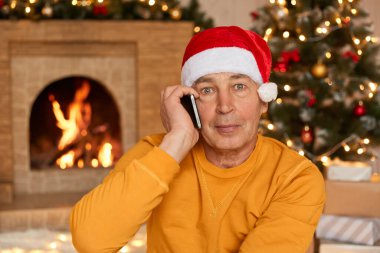 Noel Baba 'nın kırmızı şapkalı yakışıklı adam şömine ve Noel ağacının yanında otururken telefonla konuşuyor, kameraya ciddi ciddi bakıyor, sarı günlük gömlekler giyiyor..
