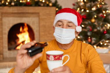 Noel Baba şapkalı hasta bir adam, şöminenin yanında oturmuş, elinde haplarla Noel süslemeleri ile festival odasında poz veriyor, Noel Baba şapkalı hasta olgun bir erkek..