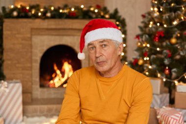 Son sınıf öğrencisi Noel Baba ve sarı kazak giyer, kameraya düşünceli bir şekilde bakar, arka planda xmas ağacı ve şömineyle poz verir, yeni yıl arifesini evde kutlar..
