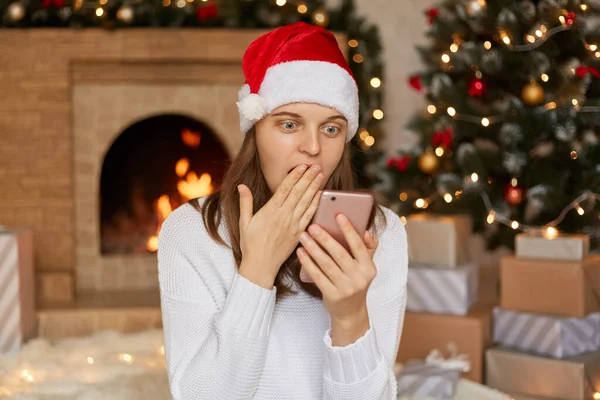 Amazed woman reading phone content in christmas hat, sees something astonishment, covers mouth with palm, looks at device\'s screen with big eyes, posing in living room with fireplace and x-mas tree.