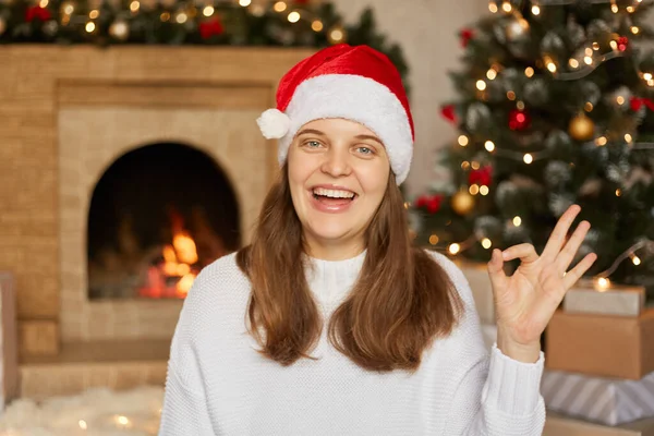 兴奋的年轻女子头戴圣诞帽 身穿套头衫 摆出一副不错的手势 兴奋地看着相机 露出牙齿 站在室内 背对着壁炉和圣诞树 — 图库照片