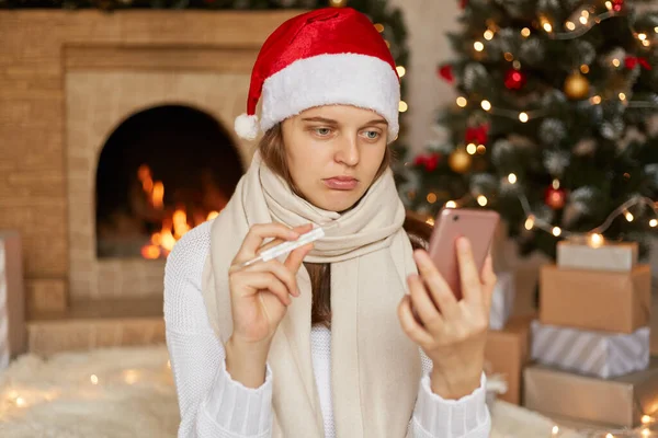 Young woman wearing Christmas hat, take video calls during corona virus pandemic, with Christmas tree and fireplace on background, showing thermometer to camera, telling about her high temperature.
