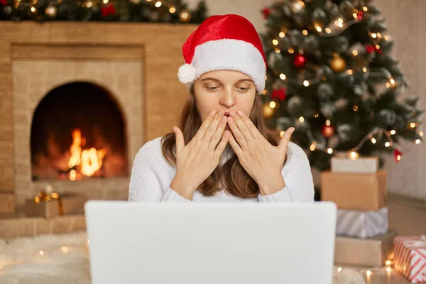 Happy woman talking via video call with somebody, greeting closer people with Christmas holidays, blowing air kisses, sitting in living room near x-mas tree and fireplace.