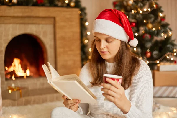 女性は熱い飲み物と暖炉とクリスマスツリーの近くの本のカップで休んで ページに集中して見て 余暇時間を過ごして お祝いの帽子と白いニットセーターを着て — ストック写真