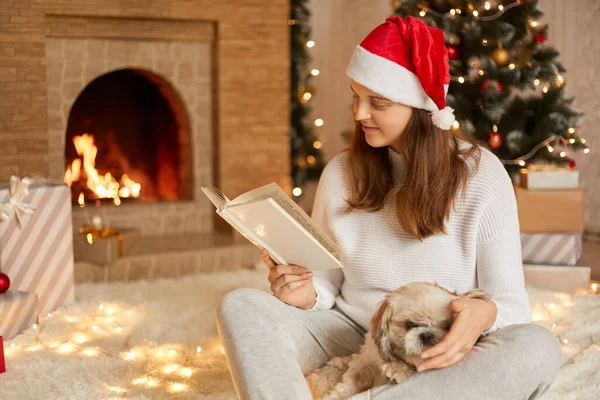 積極的な若い女性楽しい時間を過ごすために彼女の犬 かわいい女性ポージングでお祝いのリビングルームで彼女のペット 読書本ながらクリスマスツリーと暖炉の近くに座って — ストック写真