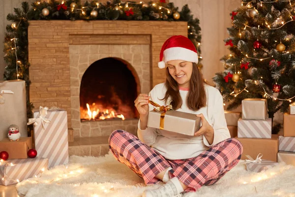 お祝いのリビングルームで柔らかいカーペットの上に座っている間 白人女性は現在のボックスを開き 女性は家でクリスマスを祝う チェッカーパンツ 白いシャツとサンタクラスの帽子を着て — ストック写真