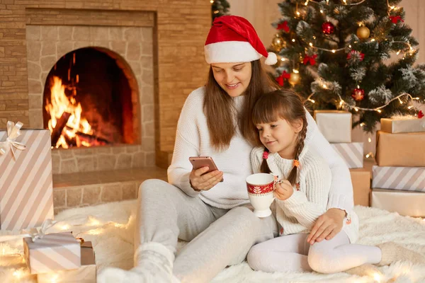 Happy mother and little daughter with smart phone at home sitting on floor, looking at device screen, having video call, posing in living room with Christmas decoration.