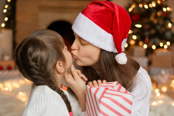 Girl surprising her mother with Christmas gift, mommy kissing her child with closed eyes, woman wearing red santa claus hat, being photographed in room with New Year decoration.