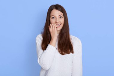 Parmaklarını ısıran ve doğrudan kameraya bakan güzel bir kadının portresi mutlu görünüyor, beyaz bir gömlek giyiyor, mavi duvara yaslanmış, siyah saçlı..