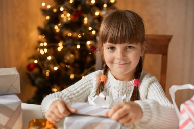 Oturma odasında masada otururken hediye kutusunu toplayan kız, büyüleyici bir gülümsemeyle kameraya bakıyor, örgülü saçlar takıyor, beyaz kazak giyiyor, arka planda xmas ağacı ile poz veriyor..