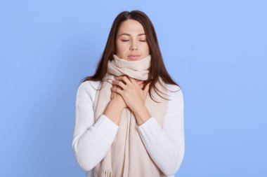 Beyaz gömlekli, atkıya sarılmış, boğazı ağrıyan, elini boynunda tutan genç bir kadın. Boğaz ağrısı, acı verici yutkunma. Koyu saçlı kadın, mavi arka planda yalnız poz veriyor..