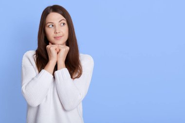 Güzel, düşünceli, genç Avrupalı bir bayan elini çenesinin altında tutuyor, sağlıklı bir teni var, beyaz bir kazak giyiyor, düşünceli görünüyor, bir şey hakkında düşünüyor, mavi duvarın arkasında duruyor. Boşluğu kopyala.