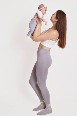 Neşeli, güzel, genç bir kadın kucağında bebeğiyle ona sevgiyle bakıyor. Beyaz stüdyo arka planında izole bir şekilde poz veriyor. Gri tayt ve sutyen giyen bir kadın..