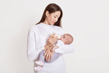 Anne şişeden besleniyor, bebeği beyaz arka planda izole edilmiş, kahverengi saçlı, süveter giyen, kucağında bebek olan, çocuklu genç anneye bakıyor..