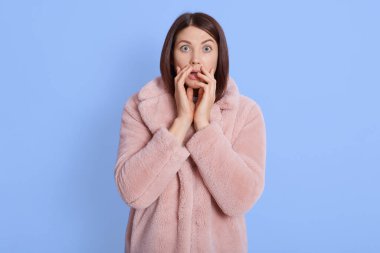 Şok olmuş genç bir kadın, mavi arka planda tek başına dikiliyor, büyük gözlerle kameraya bakıyor ve soluk pembe kürk manto giyiyor, şaşırmış yüz ifadesi takınıyor..