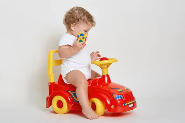 Little blond driver sitting on red tolokar, wearing white bodysuit, posing barefoot, holding tiny car in hands, looking concentrated on his toys, having wavy hair, kid playing indoor.