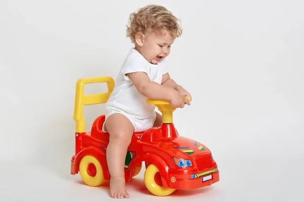 Funny kid wearing white bodysuit posing barefoot isolated over white background, being upset, starts crying, playing with red toy car, child driving toy vehicle.