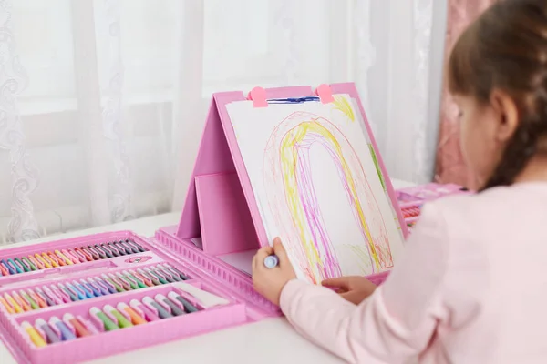 Talented child girl draws rainbow with colored pencils while sitting at table with set of art and creativity, has lots colors of wax crayons, making illustration.