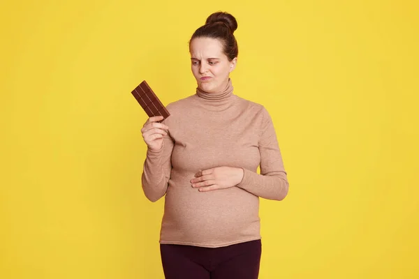 若い女性の疑いがチョコレートを食べるかどうか 黄色の背景に隔離されたポーズ 片手でチョコレートのバーを保持し 疑いの顔の表情で見て 健康的な食事を維持する必要があります — ストック写真