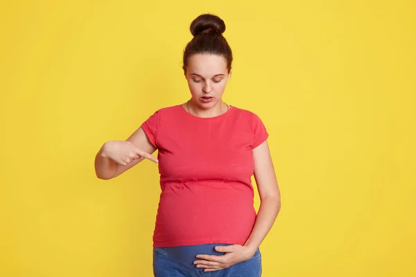 彼女の腹を見て それを見て 彼女のおなかの上にインデックス指で指して 赤いTシャツを着て 黄色の壁の上に隔離されたスタンド — ストック写真