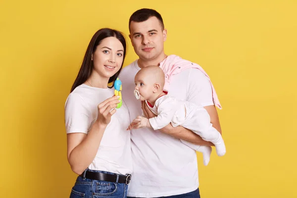 父と赤ちゃんの子供は黄色の背景に隔離されたポーズ 手にビーンズバッグを持つママ 家族はカジュアルな白いTシャツを着ています 生まれたばかりの娘を持つ幸せな両親 — ストック写真