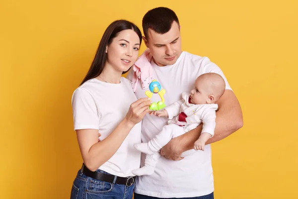 有孩子的年轻父母 黑头发的妈妈 身穿白色T恤 给孩子们展示玩具 父亲抱着小女儿 家人在黄色背景下孤立无援 — 图库照片