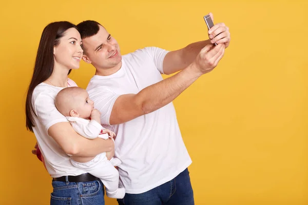 黄色の背景に隔離された携帯電話で自撮りしている赤ちゃんを持つ家族 スマートフォンで写真を撮る白いTシャツを着たカップル デバイスカメラを見て笑顔 — ストック写真