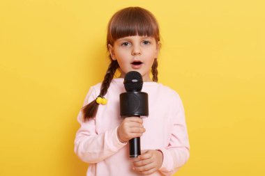 Küçük Avrupalı kız çocuğu mikrofonda şarkı söylüyor, konsantre görünüyor, harika bir şarkıcı olmak istiyor, küçük sevimli bir sanatçı konseri ayarlıyor, soluk pembe süveter giyiyor..